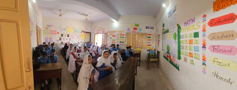 A classroom with students seated at wooden desks and colourful maths charts on the walls, including a Fraction Wall and Math Symbols.