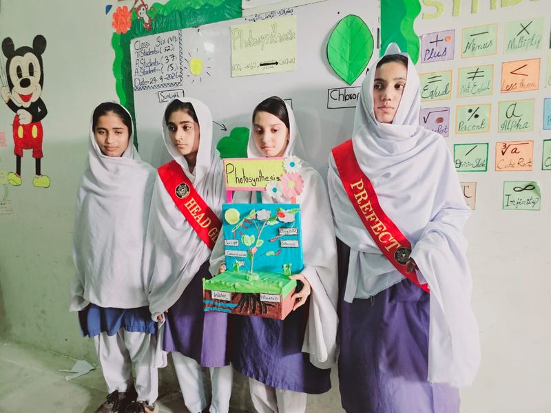 Four students stand in front of a photosynthesis chart, holding a model of a plant with sunlight, water and minerals labelled. Two wear red sashes.